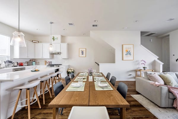 Open-plan kitchen, dining, and living area with white cabinetry, wood dining table set for six, and gray sofa with staircase beyond.