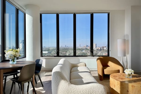 Modern living room with floor-to-ceiling windows overlooking city skyline, cream sofa, mustard armchair, and wooden dining table.